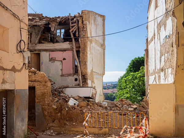 Obraz Vista de un edificio en ruinas con una valla amarilla de aviso, piedras  y restos de construcción con peligro de derrumbe. Verano de 2021 España.