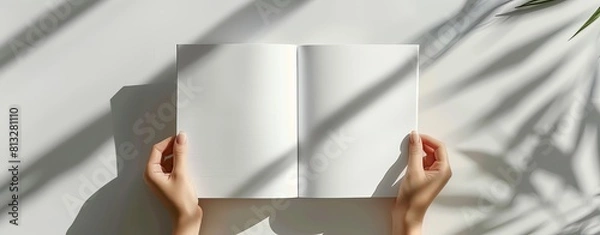 Obraz A woman's hands open an empty white book mockup on a light grey background, in a flat lay with shadows from a window, in the style of a modern minimalist. A4 format for presentation of magazine