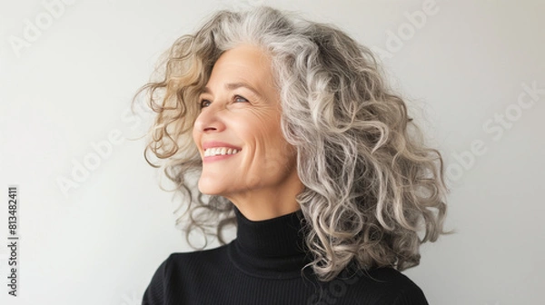 Obraz Middle-aged woman with curly silver and gray hair smiles in side view against white background.
