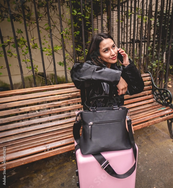 Obraz Stylish and young traveler latina brunette with fashion look and pink suitcase exploring city park while using the phone