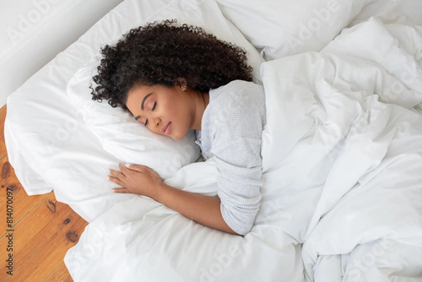 Fototapeta Hispanic woman is lying down on a bed with a white comforter covering her, appearing relaxed and comfortable in a bedroom setting, view above