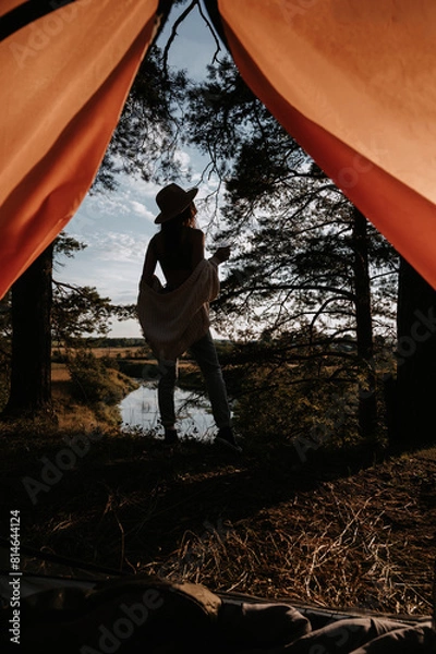 Fototapeta Camping. View from the tent. The silhouette of a young woman standing at the edge of a cliff against the background of trees and water. Outdoor recreation.