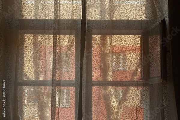 Fototapeta A window with a white wood frame overlooking the neighboring house, sun is shining through thin translucent curtains.