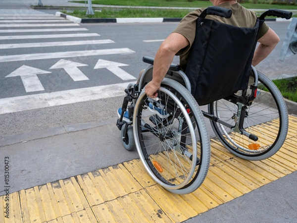 Fototapeta Rear view of an elderly woman in a wheelchair going to a pedestrian crossing. Close-up on wheels. 