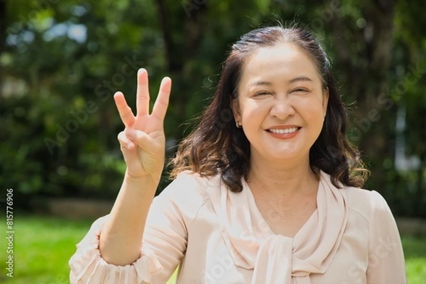 Fototapeta Healthy happy smiling middle aged asian woman pointing 3 fingers up, concept of three points, number three, third count in summer green park