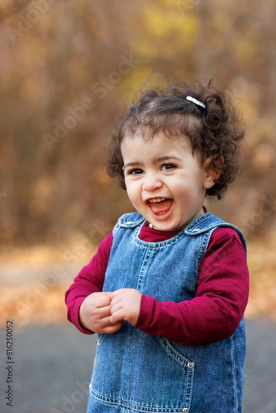 Fototapeta Cute laughing kid in overalls and red top stands still in a world of autumnal hues