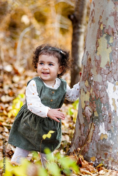 Fototapeta Smiling little girl standing by a tree, touching its bark, leaves at their feet