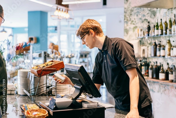 Fototapeta Young waiter serving customer at cash point in cafe. Man working with POS terminal. Cashier, barista checking for payment receipt. Hospitality, server and preparing a slip at the till in coffee shop.