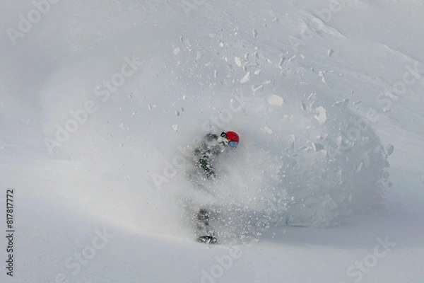 Fototapeta A snowboarder rides out of a cloud of snow breaking in pieces after performing a powder turn on an untouched slope