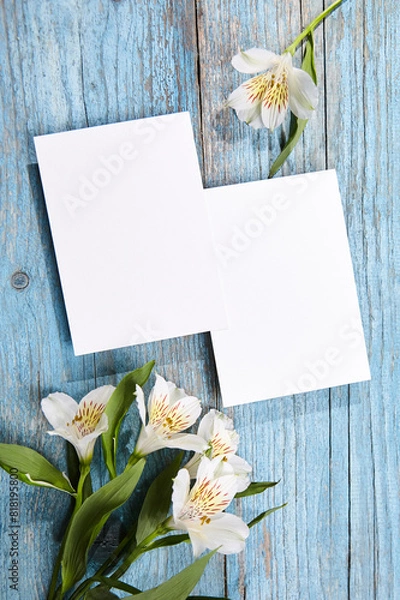 Fototapeta Two blank cards in portrait orientation placed on a blue wooden surface, accompanied by a cluster of white Alstroemeria flowers