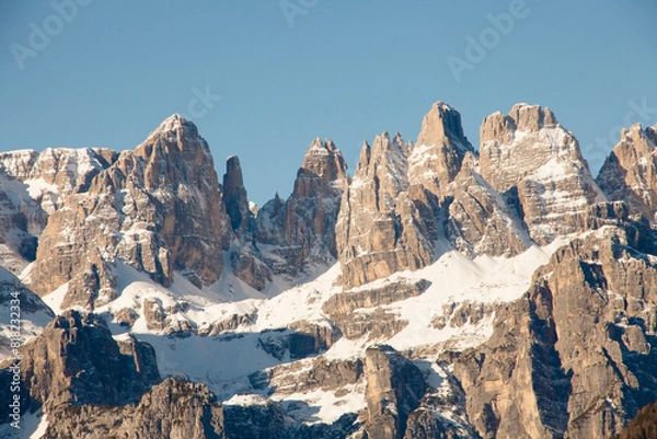 Fototapeta The stunning mountain Paganella in Italy, with rugged, jagged peaks partially covered in snow. Steep rock faces and snow-dusted ridges. Majestic landscape under a clear blue sky.
