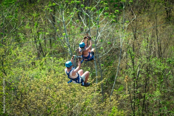 Fototapeta A couple riding a zipline in Costa Rica while on vacation. View from behind as the tourists zip down a jungle mountain on an adventure tour.