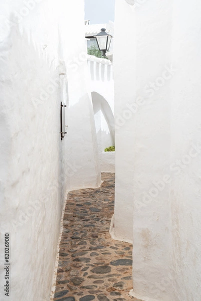 Fototapeta A picturesque view of an empty street in Binibeca, Menorca. The whitewashed buildings and cobblestone pathway showcase the charming Mediterranean architecture of this tranquil village