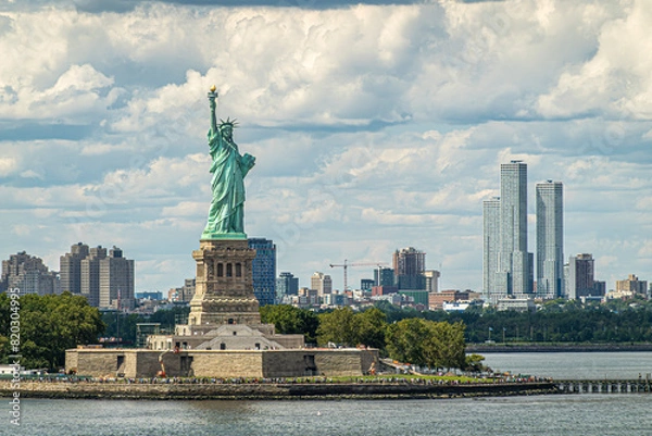 Fototapeta New York, NY, USA - August 1, 2023: Jouned Squared tower of Jersey city is seen to the right of Statue of Liberty on its island under blue cloudscape. Plenty of visitors and more building in back