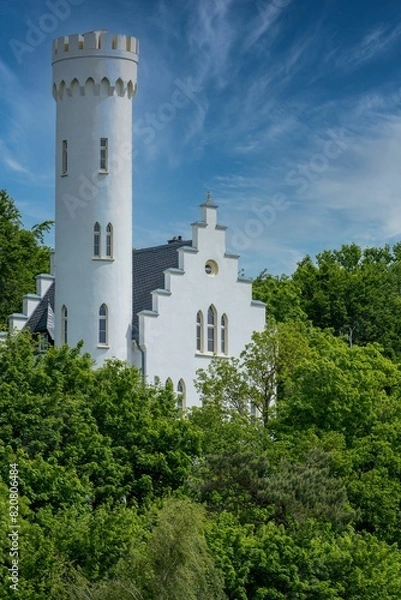 Fototapeta Majestic white tower standing tall against a backdrop of lush green trees and a bright blue sky