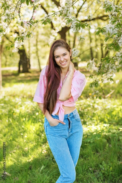 Obraz Spring.Beautiful laughing girl in a pink shirt and jeans standing in a blooming garden. Young model in a great mood between the apple trees.