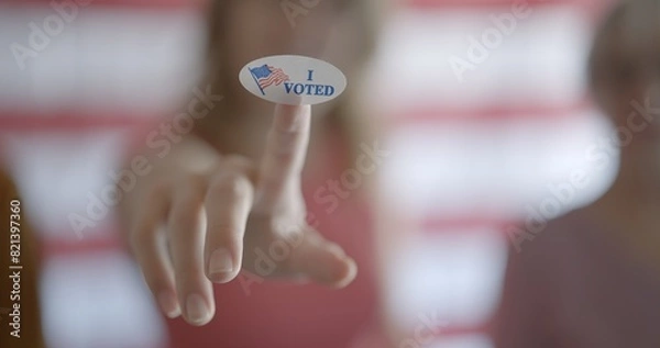 Fototapeta Young woman holds up oval "I voted" sticker on finger, pushed towards camera. Shallow depth of field with focus on sticker and people blurred