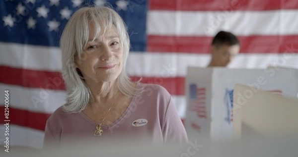Fototapeta Elderly woman voter wears "I voted" sticker and smiles to camera. Asian voter, soft focus, in booth behind her. US flag on back wall