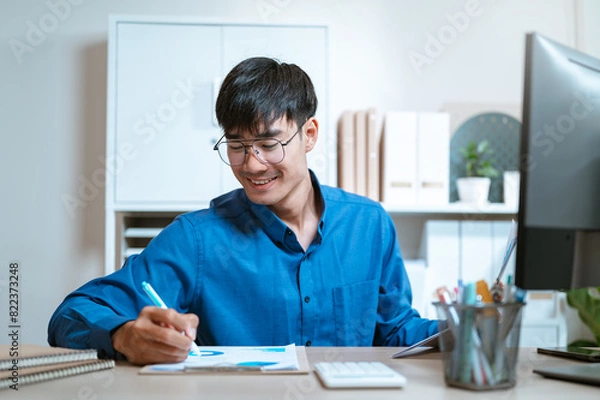 Fototapeta document, thinking, working, writing, executive, expertise, ideas, planning, job, management. A man is sitting with a blue shirt on and writing with a blue pen. He is smiling and enjoying himself.