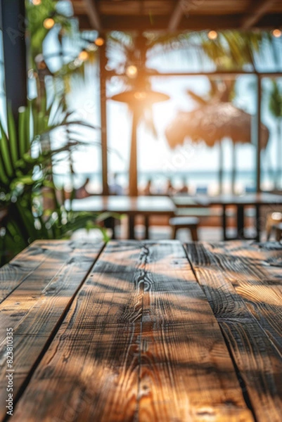 Fototapeta A wooden table in the foreground with a blurred background of a beachside cafe. The background shows sandy beach views through large windows, casual seating, tropical plants, and customers
