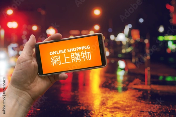 Fototapeta Online shopping, China import visualization. Hand holding a phone with orange text "online shopping" representating a store market app. Same in Chinese language. Colorful city lights in background. 