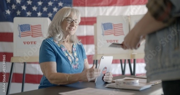 Fototapeta Three quarter angle, medium shot, elderly woman election worker checks voter identification for young female voter using tablet computer at polling station. Flag and voting booths behind