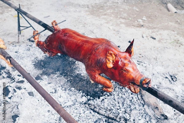 Fototapeta Closeup of the popular and delicious roasted pig known as "lechon baboy" skewered on a spit over burning charcoals on slow cooking or grilling outdoor. Selective focus.