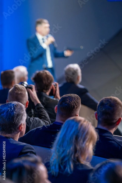 Obraz Speaker is addressing an audience at a business conference. People in the audience are paying attention and some are taking photos.