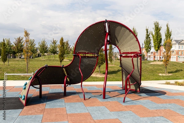 Fototapeta Modern playground equipment - red and grey - set on colorful rubber tiles - suburban homes and greenery in background. Taken in Toronto, Canada.