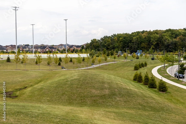Fototapeta Suburban park with green rolling hills - paved walkways and young trees - playground equipment - residential houses background - tall light poles and partly cloudy sky. Taken in Toronto, Canada.