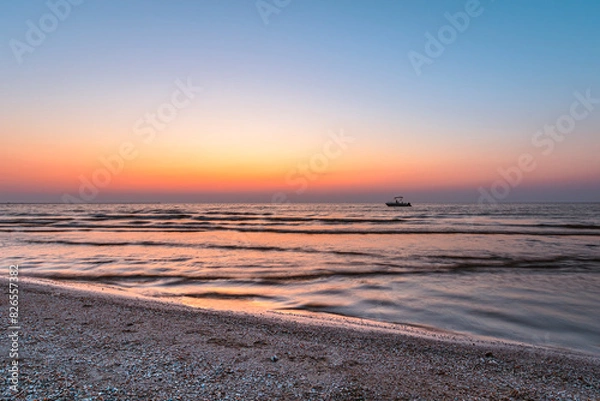 Fototapeta A serene sunset over a calm sea with gentle waves lapping at a pebbled beach. A small boat is silhouetted against the vibrant hues of the sky, creating a tranquil and picturesque scene perfect for con