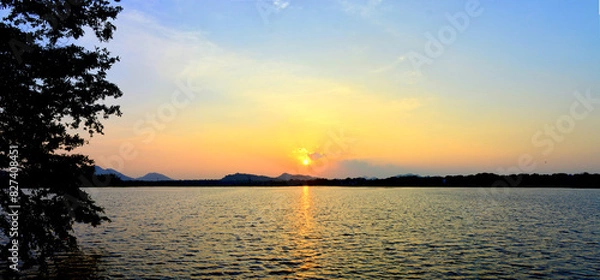 Fototapeta Panoramic view of a golden evening sunset above a natural lake with mountains silhouette backdrop