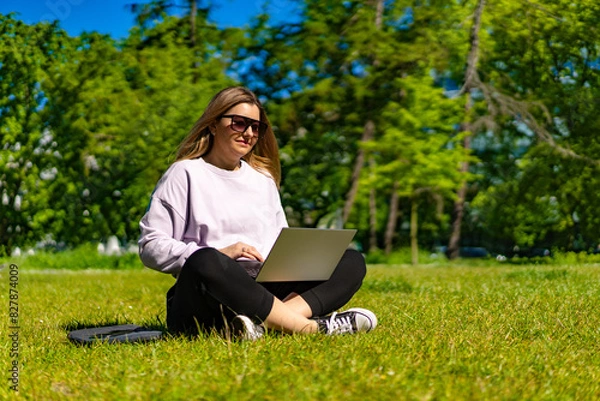 Fototapeta Beautiful woman using laptop sitting on grass in city park
