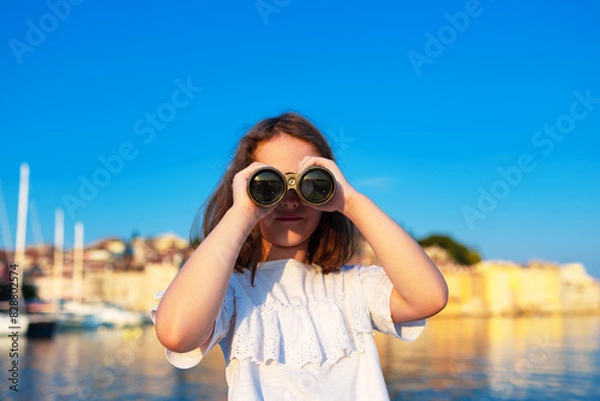 Fototapeta Funny little girl looking through vintage binoculars on sunny summer day. Rovinj town in background. Travel and adventure concept.
