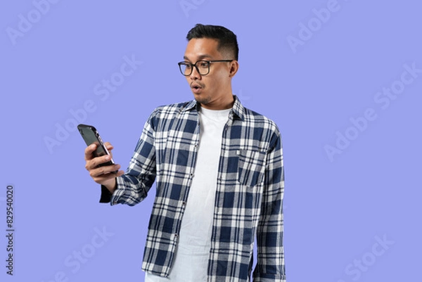 Fototapeta Portrait of a young cheerful excited Asian man standing isolated over a purple background, using a mobile phone