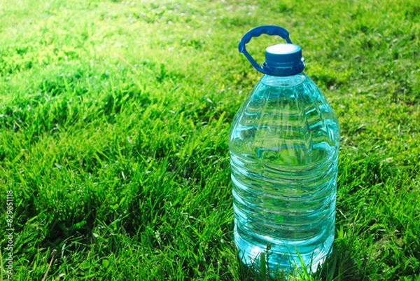 Obraz Big plastic bottle with water on the green grass background on sunny day. Close-up of water bottle and cap