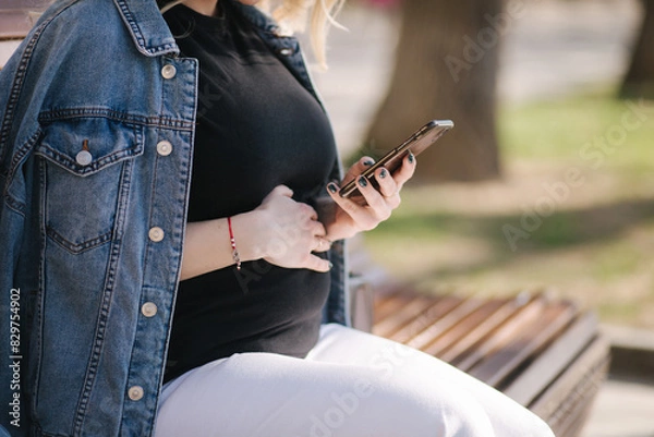 Fototapeta Middle selection of pregnant woman sits on bench in the park and using phone. Happy female have rest outdoor during the walks