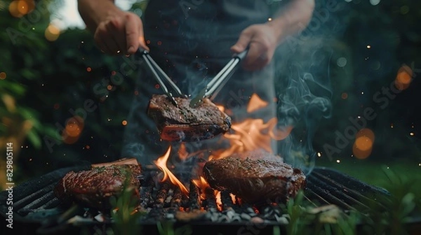 Fototapeta Close up of hands with tongs grilling marinated beef steaks on fire in the garden, outdoorsy scene. A person is using metaltipped food buner to light flames under an outdoor grill where meat.