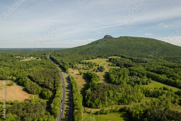 Fototapeta Highway Winds Around Green Forest that Surrounds Mountain Peak Under a Blue Sky