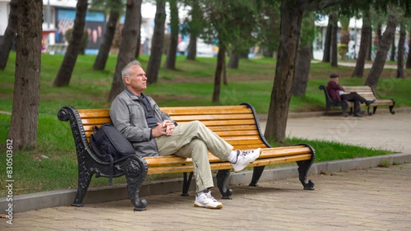 Fototapeta An elderly man with a backpack is sitting on a bench in a city park
