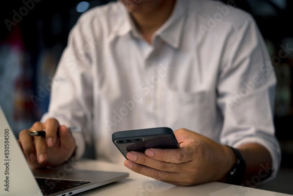 Fototapeta Businessman holding smartphone to communicate work with digital internet, online technology and laptop computer at work desk