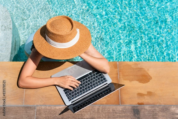 Fototapeta Young Asian traveler woman working on computer at pool in luxury resort during her summer holiday. Work from anywhere, Technology and lifestyle concept, copy space