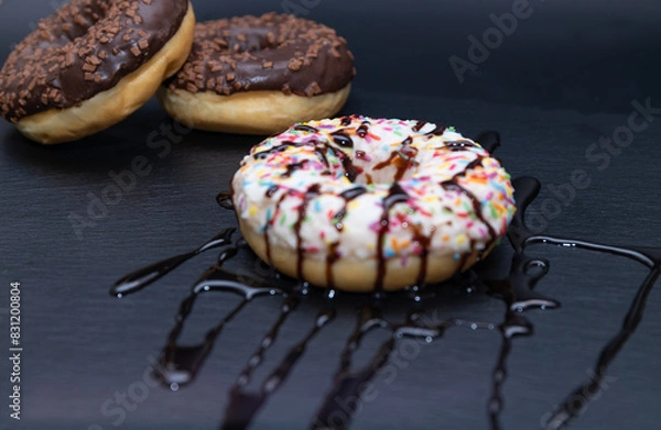 Fototapeta Sweet donuts with chocolate, sprinkled with colored sticks and doused with liquid chocolate on black plate