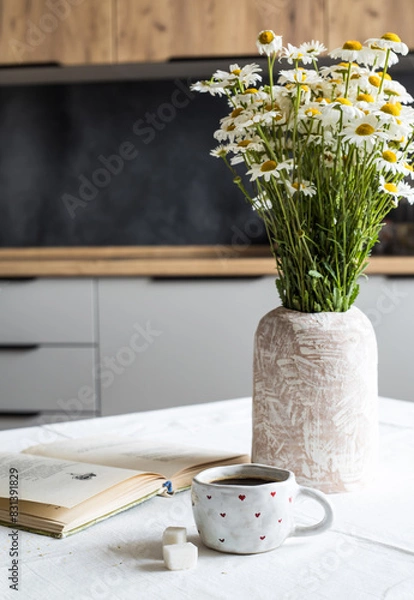 Obraz Breakfast concept. A cup of coffee, two pieces of sugar, a vase with wildflowers and an open book on a table covered with a light tablecloth against the background of the kitchen interior.