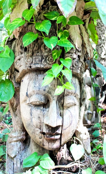 Fototapeta Buddha sculpture carved from wood, adorned with leafy vines, placed in an outdoor setting