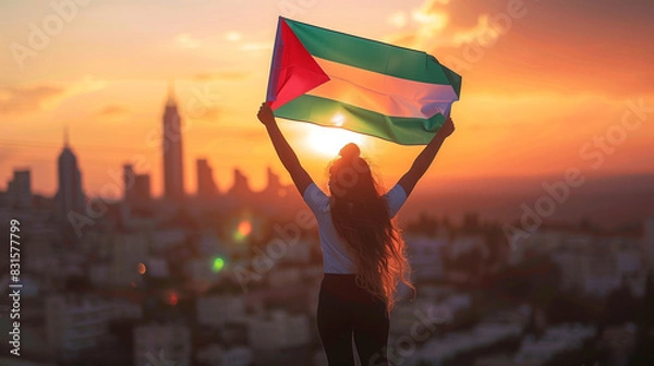Fototapeta A girl holds the Palestine flag high, with city skyline in the background at sunset. The photo is taken from behind her and she has both hands raised holding up the Palestinian Flag .