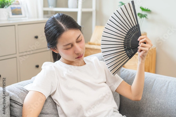 Fototapeta Suffering summer heat wave stroke, hot weather, tired asian young woman feeling discomfort sweaty, sitting on couch in living room, hand fan in blowing, wave fan to ventilation when hightemperature.