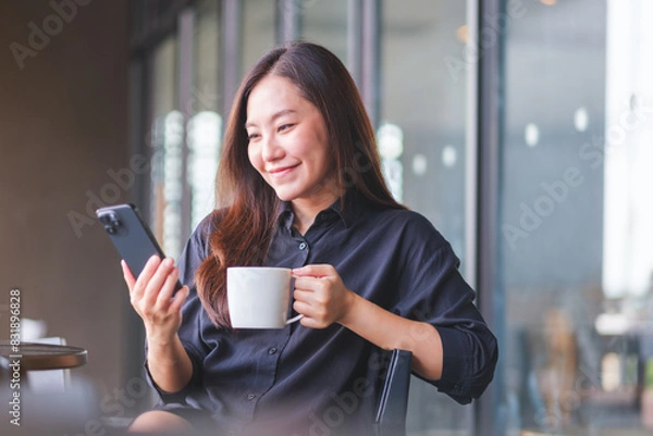 Fototapeta Portrait image of a young woman holding and using mobile phone while drinking coffee in cafe