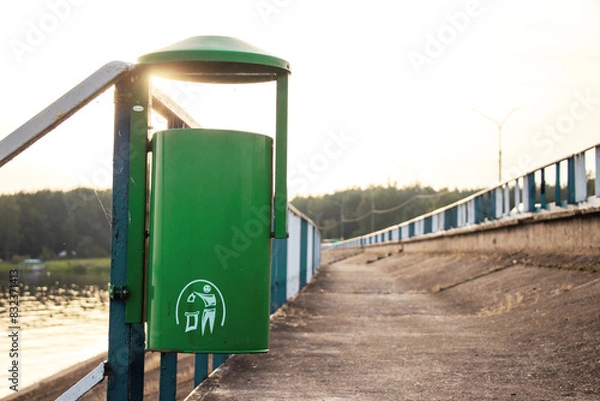 Fototapeta green color recycling bins in a public park near the river. Metal trash bin in the park with blurry sunny background and forest. Bridge and dam with street, bicycle and pedestrian path