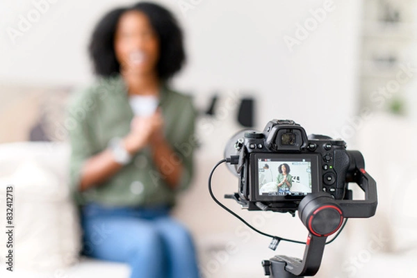 Fototapeta An African-American woman with a radiant smile is seated on a couch, interacting with a camera on a gimbal, creating content in a modern living room with soft natural light, focus on the camera